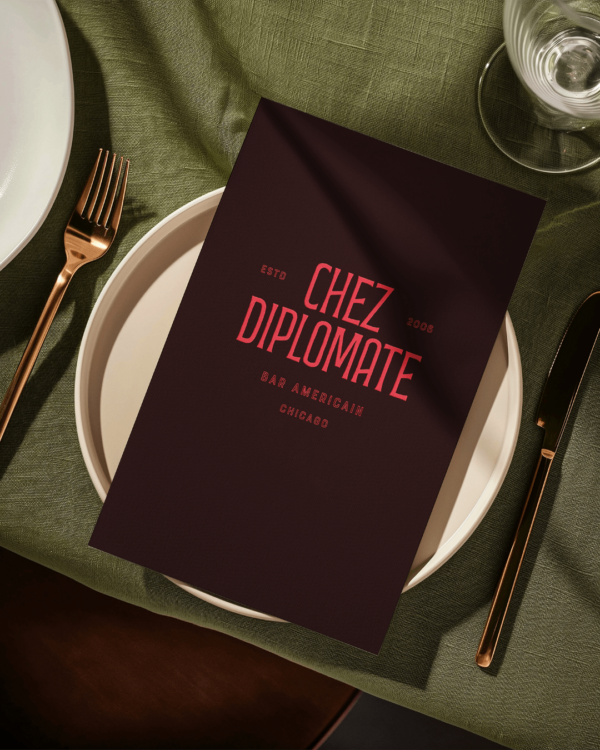 A close up of a burgandy menu book with red writing on it featuring the "Chez Diplomate" logo on front, sitting on top of a dinner plate on a deep green restaurant table.