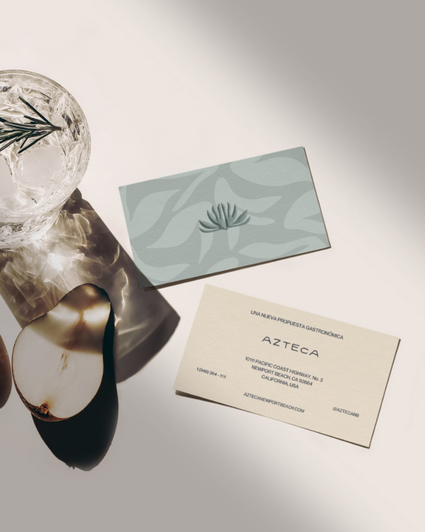 An overhead shot of two business cards (front and back design) in the Azteca brand style, sitting next to a cocktail glass and sliced pear.