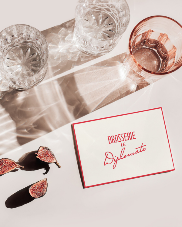 A white business card with red trim with "Brasserie le Diplomate" written on the front. Sitting on a flat surface next to several rose colored glasses and sliced figs.