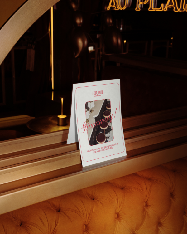 A white promo card with red writing and an image of a restaurant, perched up straight on top of an orange colored banquette and a gold-trimmed beam behind the banquette.