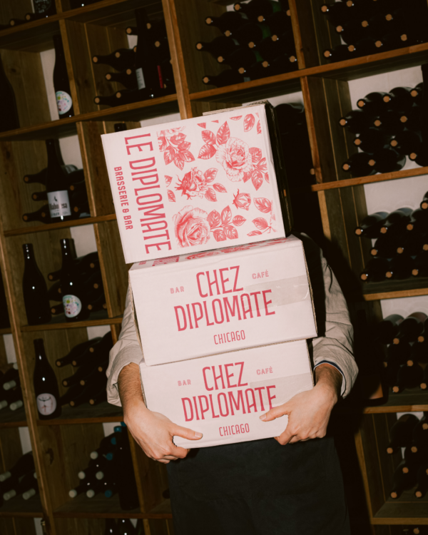 A person holding three wine boxes with racks of wine behind them. The wine boxes are pink with red flowers and red writing on them featuring the "Chez Diplomate" logo on them.