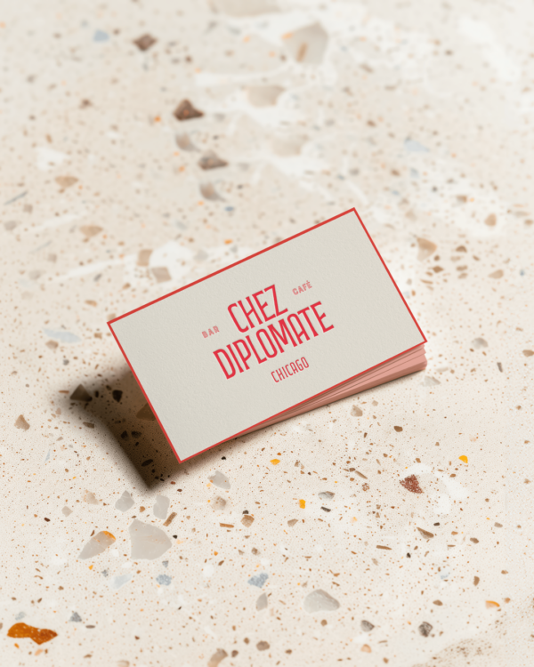 A close up of a white business card with red trim and the "Chez Diplomate" logo in red on top of a bespeckled sand colored table top surface.