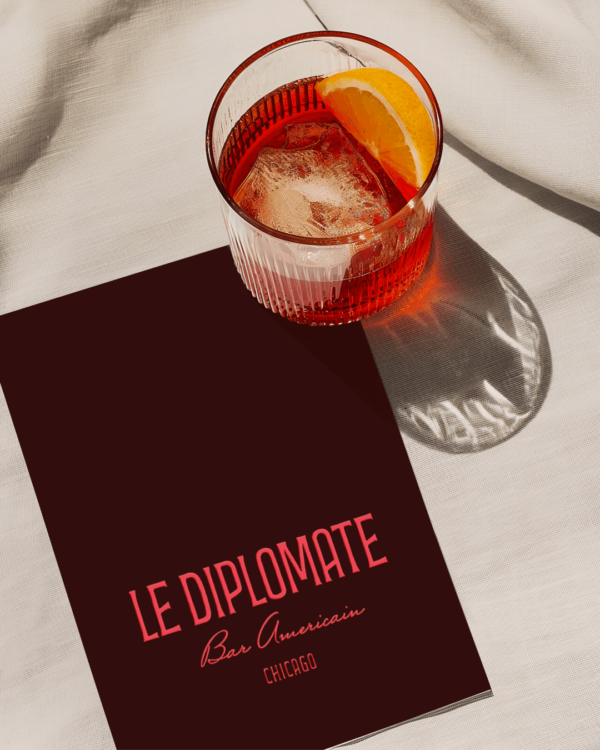 A close up of a burgandy menu book with red writing on it featuring the "Chez Diplomate" logo on front, sitting on top of a flat surface with a negroni cocktail sitting next to it.