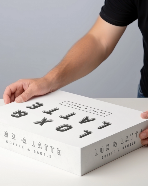 A close up of hands holding a white bakery box featuring the "lox and latte" logos and branding in black writing on the top and sides.