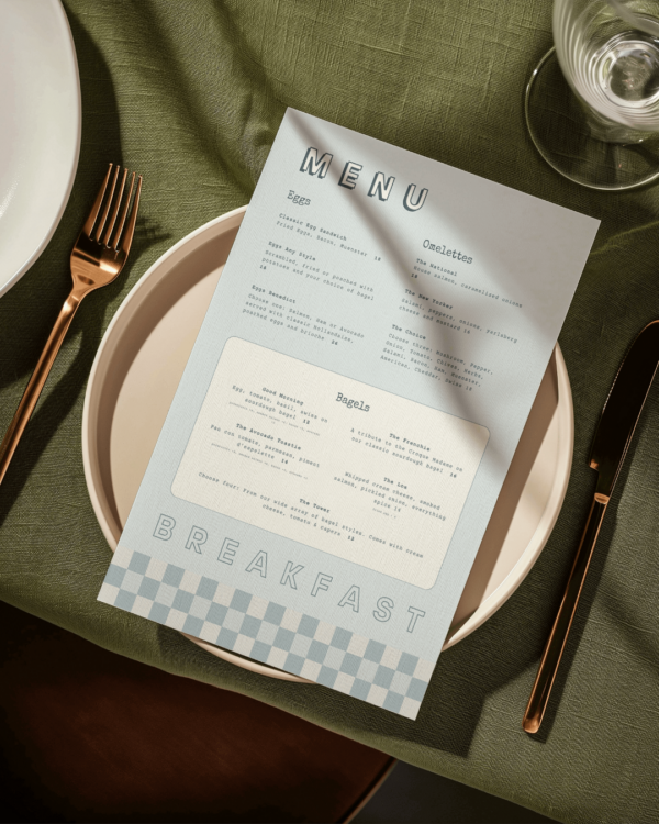 A blue and white menu sitting on top of a dinner plate on a green restaurant table with gold flatware.