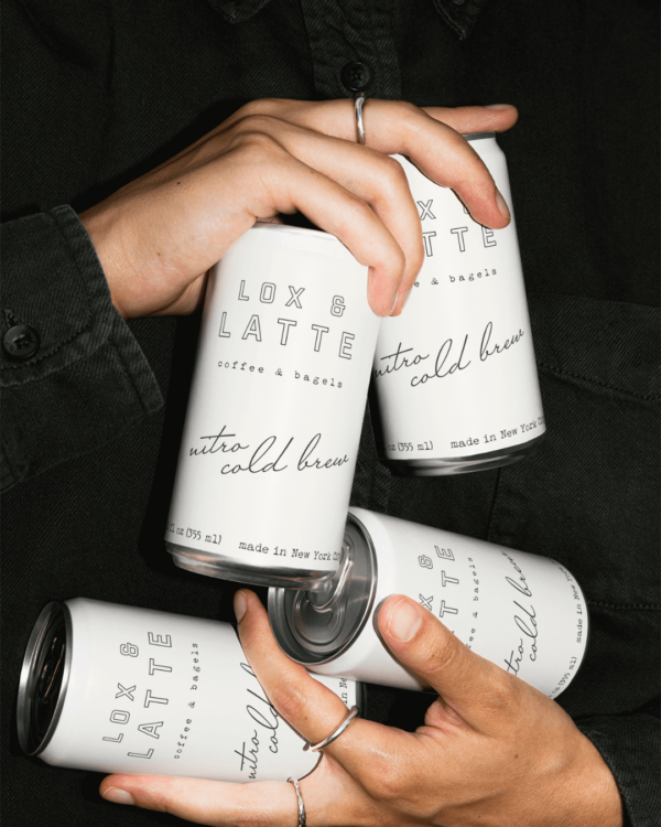 A close up of hands holding four white cans of Lox and Latte nitro cold brew featuring the Lox and Latte branding style.