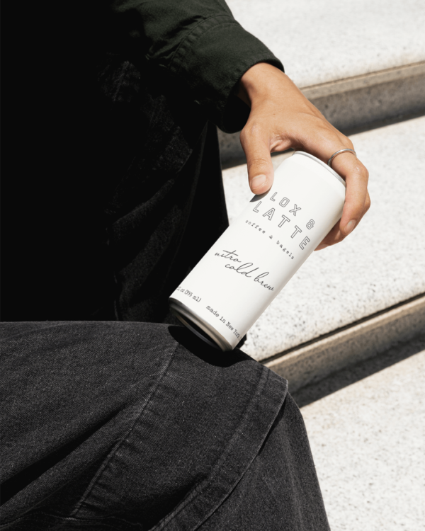 A close up of a hand holding a white can of nitro cold brew featuring the Lox and Latte logo in black writing.