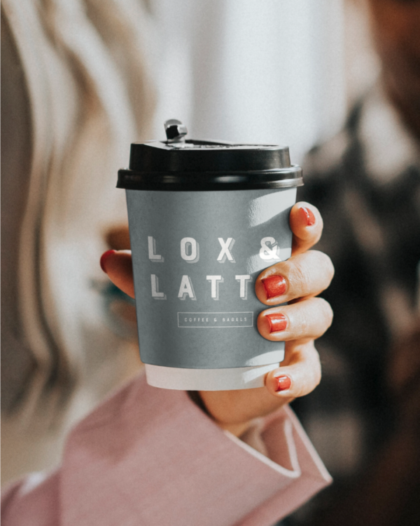 A close up of a womans hand holding a sage green coffee cup with black top featuring the "Lox and Latte" logo and branding.